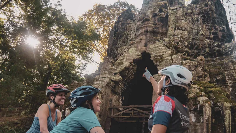 Cyclists in helmets riding toward a carved Angkor Thom gate on an Angkor temples bike tour
