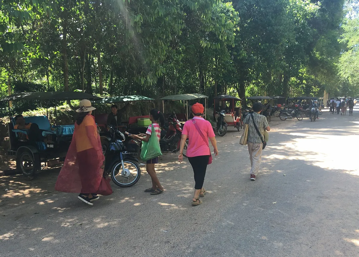 Tuk-tuks parked in the shade near Ta Prohm while visitors walk past, the way private Angkor Wat tours by tuk-tuk get around the park